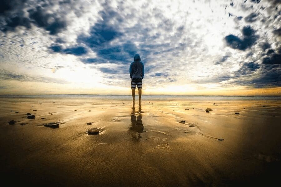 Person in a hoodie standing alone on wet sand under a dramatic sky at sunset