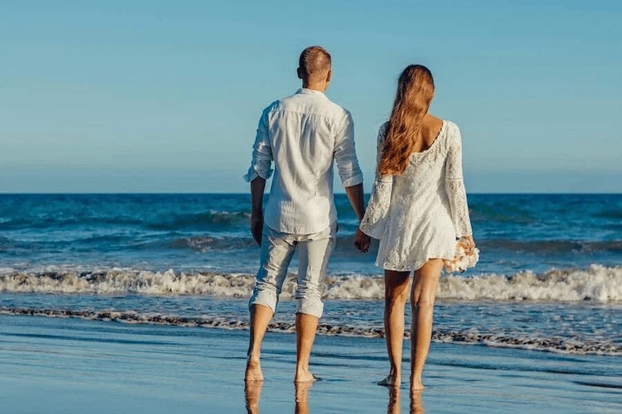 Couple in white holding hands while walking along the beach at the water's edge