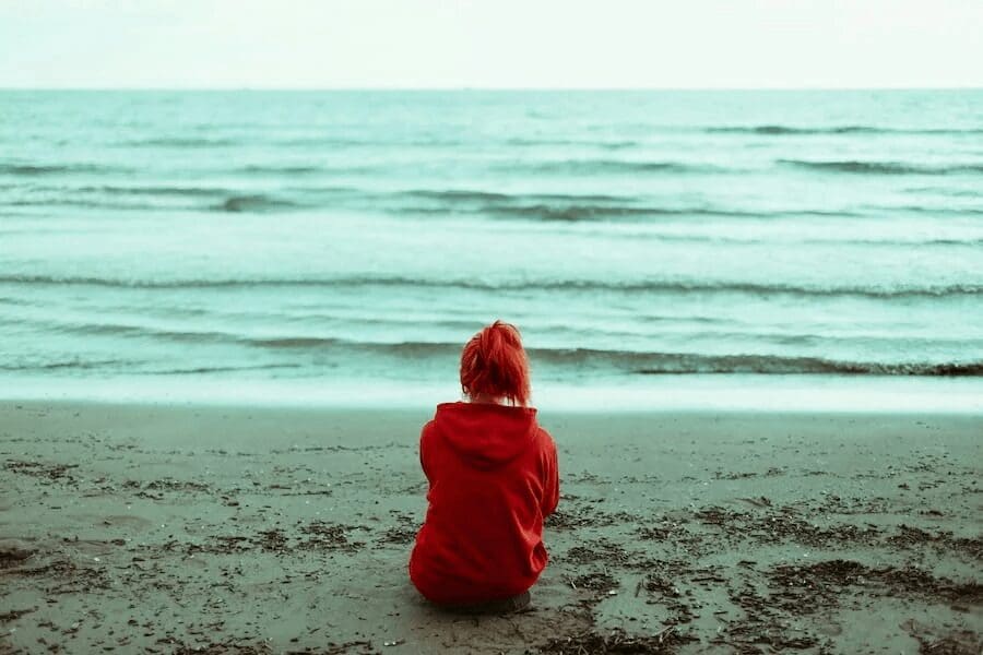 Person in a red hoodie sitting alone on the sand, facing a calm gray-green sea